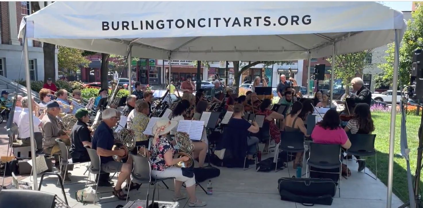 The orchestra performing outdoors in the park under a tent that says BURLINGTONCITYARTS.ORG