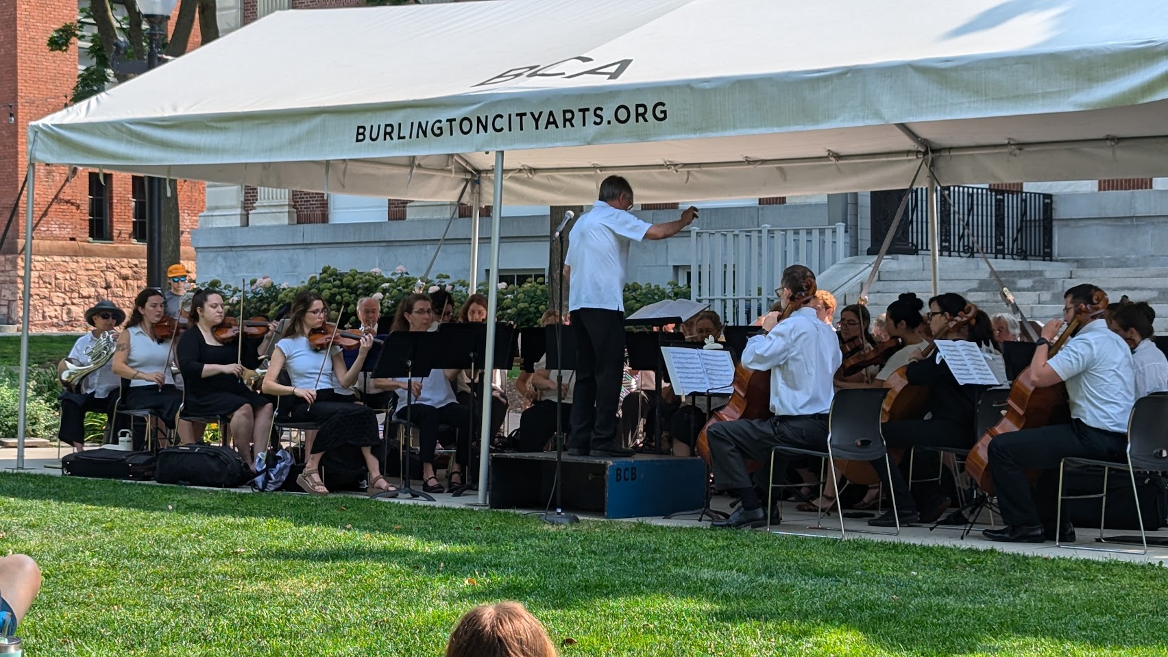 The orchestra performing outdoors in the park under a tent that says BURLINGTONCITYARTS.ORG